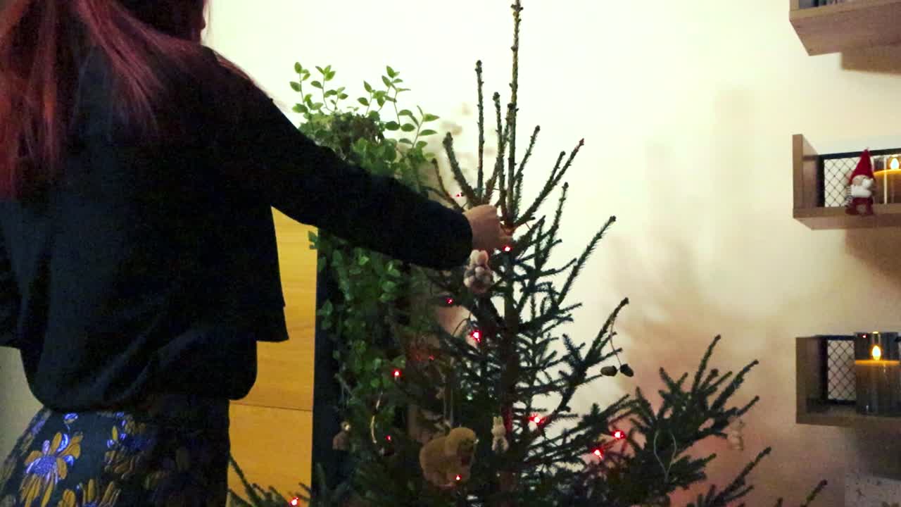 Woman with red hair, black top and a skirt placing decorations on a Christmas tree in slow motion. The Christmas tree is lit by red LED lights and candles burning on the side on the bookshelf.