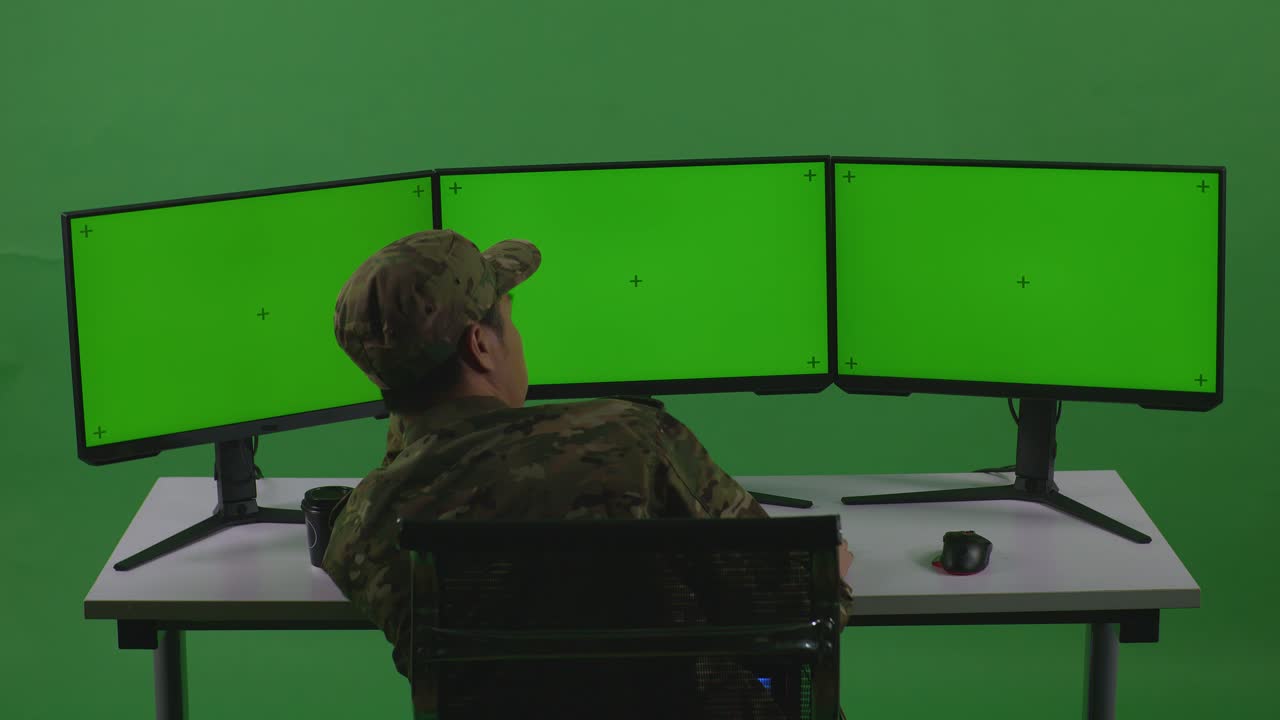 vista trasera de un soldado asiático aburrido trabajando con un simulacro de varios monitores de computadora en un estudio de pantalla verde