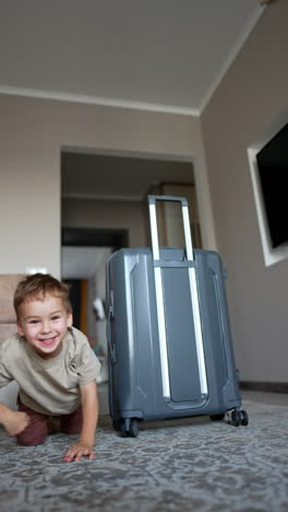 joyful caucasian toddler crawls from behind a big grey suitcase. adorable baby boy approaches camera smiling. vertical video.