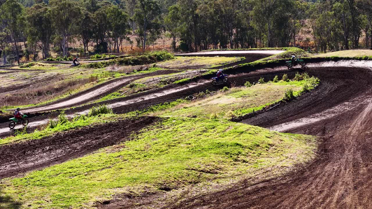 Multiple motocross riders take a dirt track curve outdoors, bright daylight, wide static shot