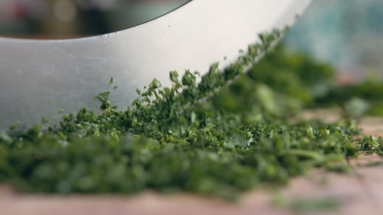 Extreme Close up front view of woman's hand fast chopping green parsley with an Italian mincing knife sharp knife on a wooden board in her kitchen. Home cooking and healthy food concept
