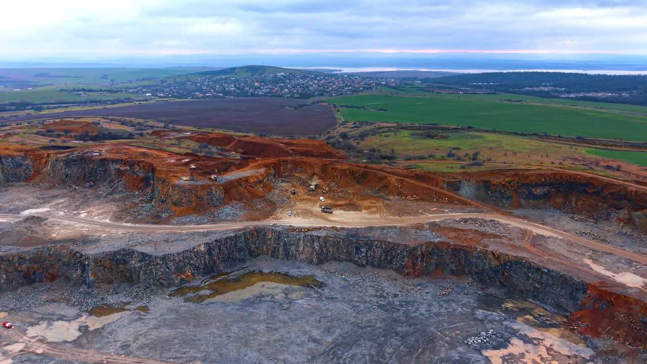 Cava a cielo aperto presso un lago. Si vede una grande cava con macchinari che lavorano sul terreno roccioso. La scena mostra campi e colline sullo sfondo.