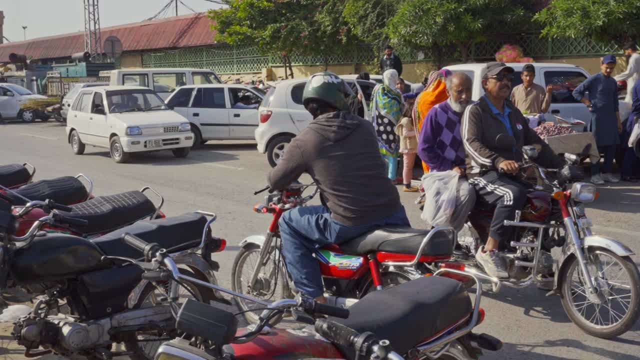 A bike taxi rider wearing a helmet patiently waits for passengers outside a bustling marketplace filled with shoppers and vendors.