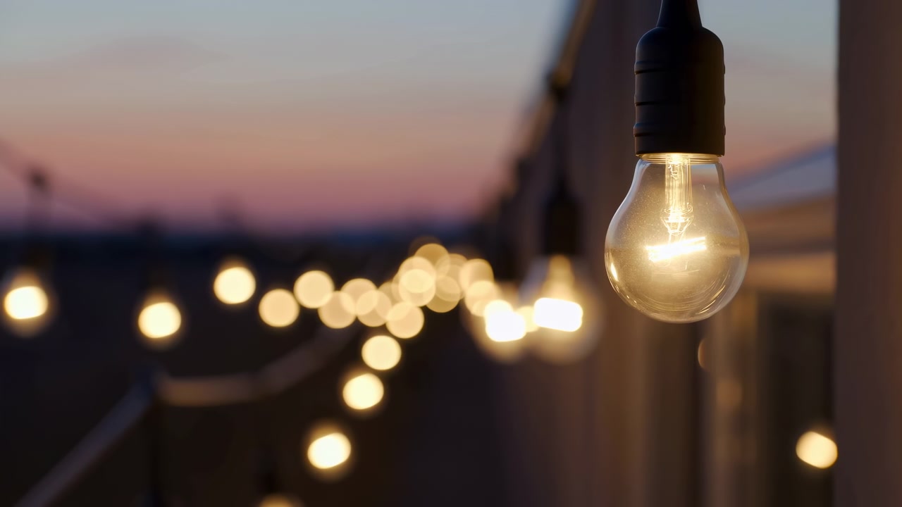 Una lampadina calda e illuminata rimane ferma vicino al bordo dell'inquadratura mentre luci decorative sfocate si perdono in profondità, con i colori del crepuscolo che svaniscono nel cielo e superfici esterne scure sullo sfondo.