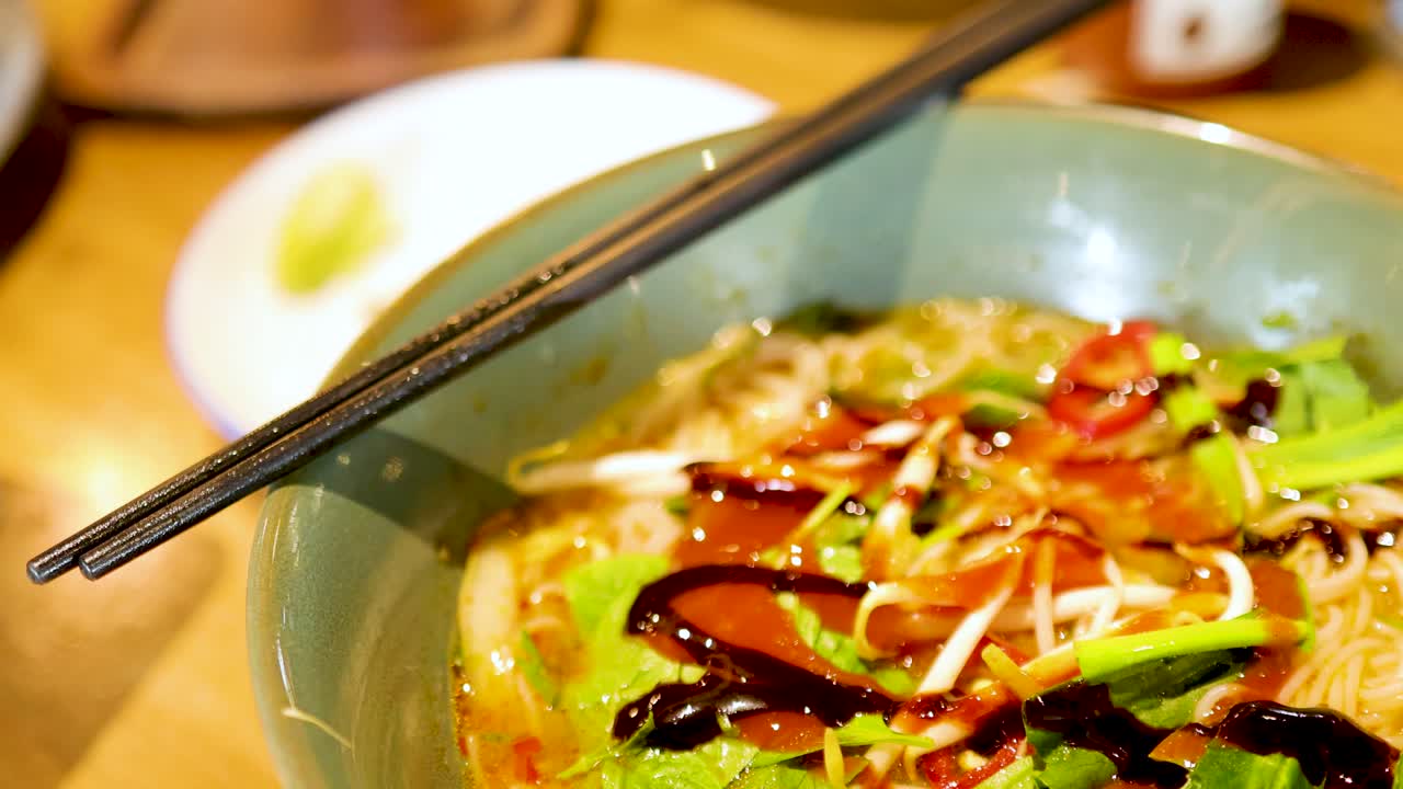 A close-up of sauce being drizzled over a vibrant bowl of Vietnamese pho, highlighting fresh ingredients and rich colors under warm lighting