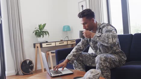 soldado masculino biracial en uniforme usando una computadora portátil y bebiendo café en la sala de estar, cámara lenta