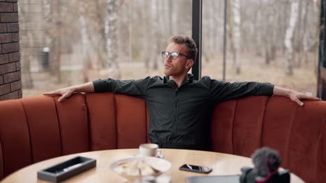 elegante hombre rizado con gafas sentado en un café reflexiona soñadoramente