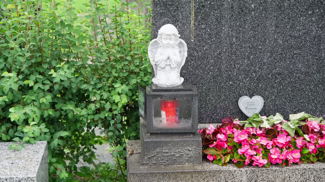 Monumento commemorativo con statua di un angelo, candela rossa e fiori rosa presso una tomba
