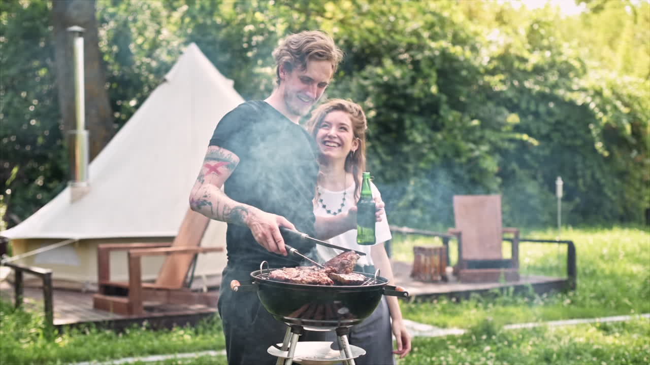 Giovane coppia che frigge carne alla griglia e beve birra, ridendo. Tanto verde intorno. Glamping. Slow motion