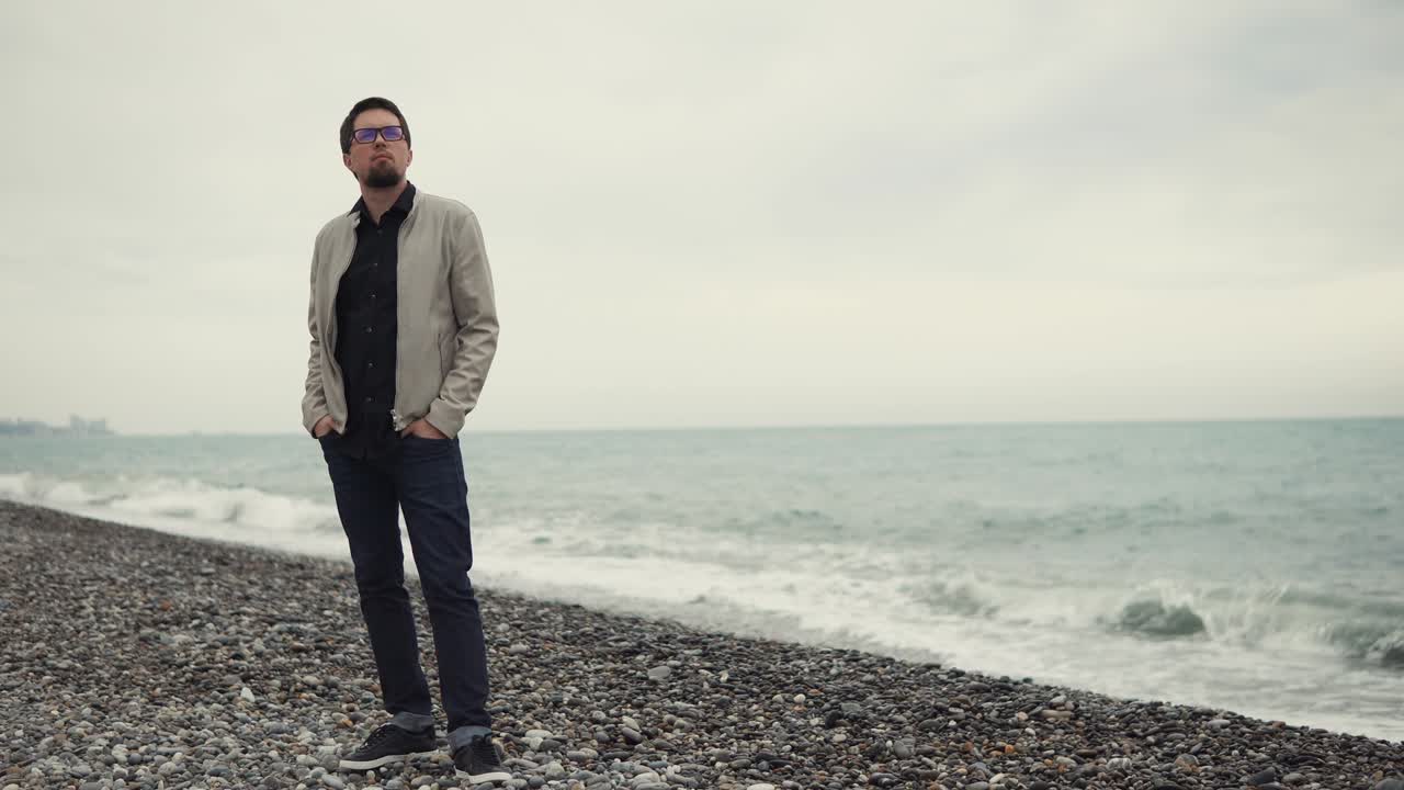a man in glasses stands on a stony beach near the sea and looks away