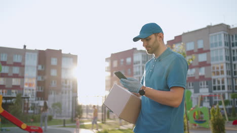 El-Cartero-Con-Gafas-Lleva-El-Paquete-Y-Mira-La-Dirección-De-Entrega-A-Través-Del-Teléfono-Móvil.-Buscar-La-Dirección-Del-Cliente-De-Entrega.-Repartidor-Con-Gorra-Y-Caja