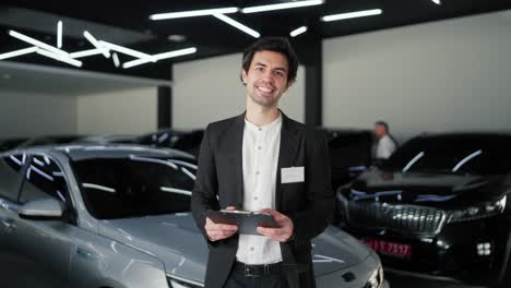 Portrait-of-a-confident-and-happy-young-brunette-guy-in-a-business-suit.-Assistant-in-a-modern-car-showroom.-A-confident-brunette-man-in-a-business-suit-jacket-and-white-shirt-stands-in-the-middle-of-a-car-dealership-with-a-tablet-in-his-hands