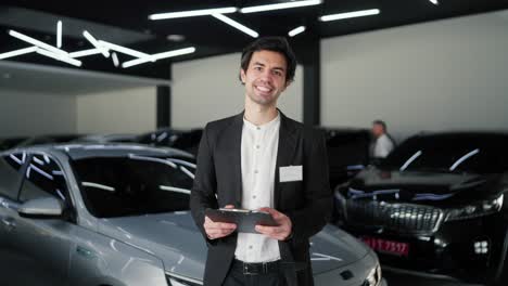 retrato de un joven moreno confiado y feliz en un traje de negocios. asistente en una sala de exhibición de automóviles modernos. un hombre moreno seguro en una chaqueta de traje de negocios y una camisa blanca se encuentra en el medio de un concesionario de automóviles con una tableta en las manos