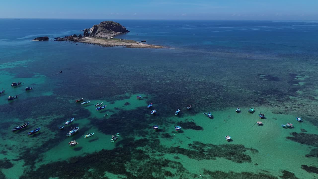 Aerial view of fishing boats navigating through shallow turquoise waters towards the island in Ninh Hải, Ninh Thuận.