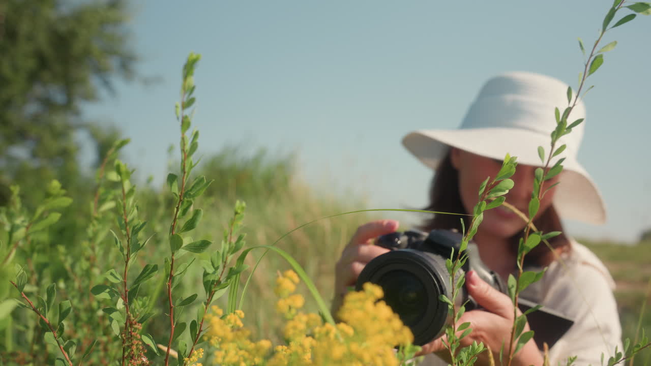 Smiling woman in white sunhat squats joyfully among green wild plants holding camera while taking photo of yellow flowers in sunny meadow surrounded by tall grasses under clear blue sky