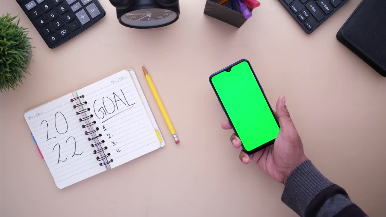 Person holding smartphone with green screen on a desk with office supplies