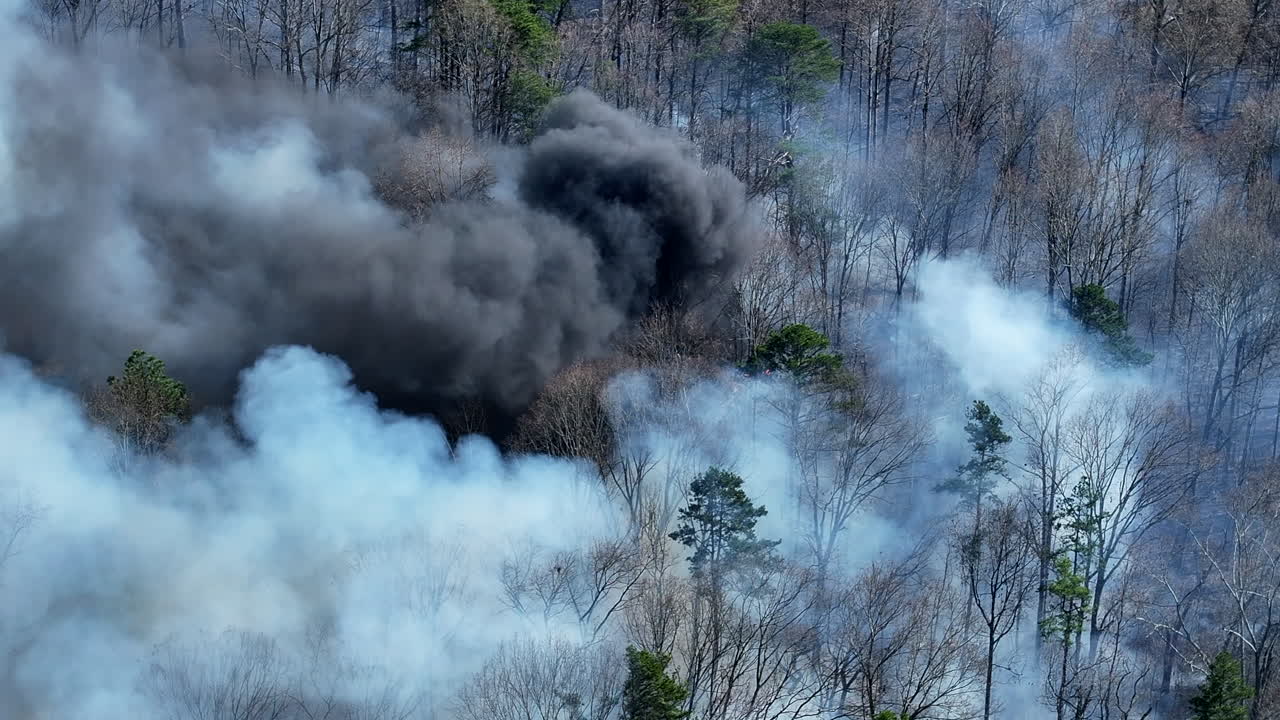 Telephoto aerial drone footage of a forest fire with gray and black smoke.