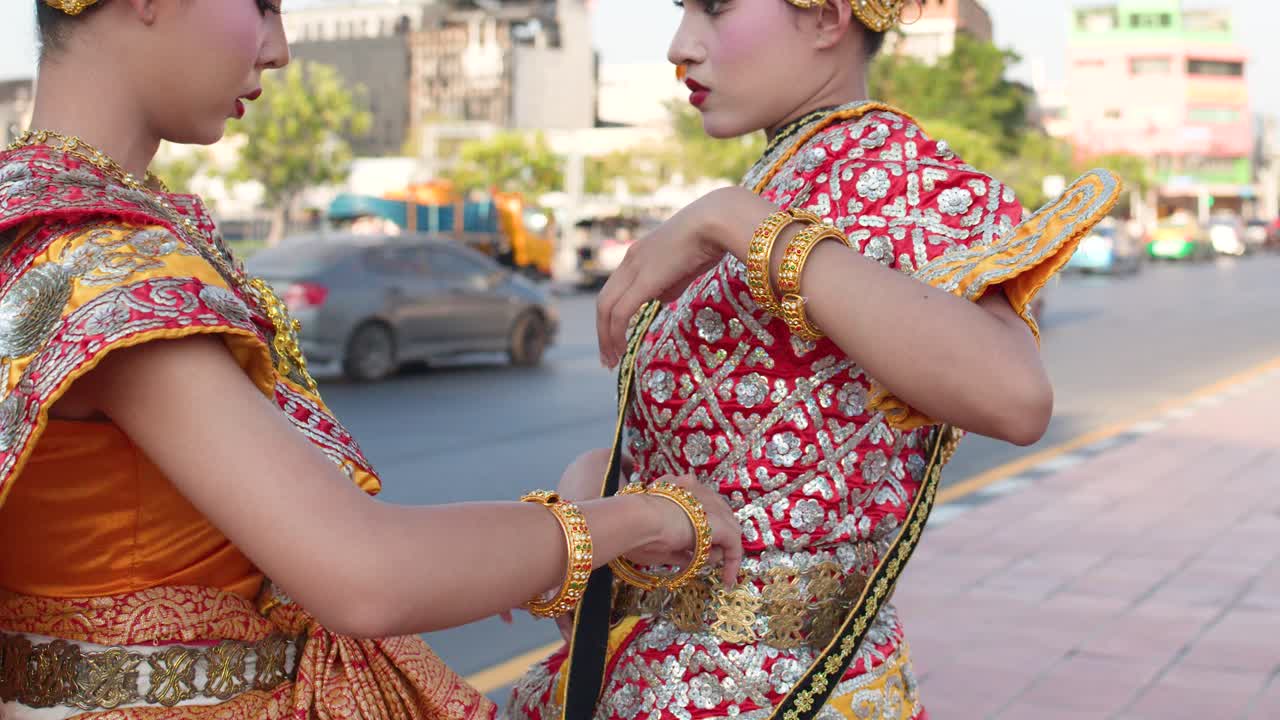 To thailandske dansere justerer udsmykkede røde og guld Khon-kostumer udendørs i skarpt dagslys