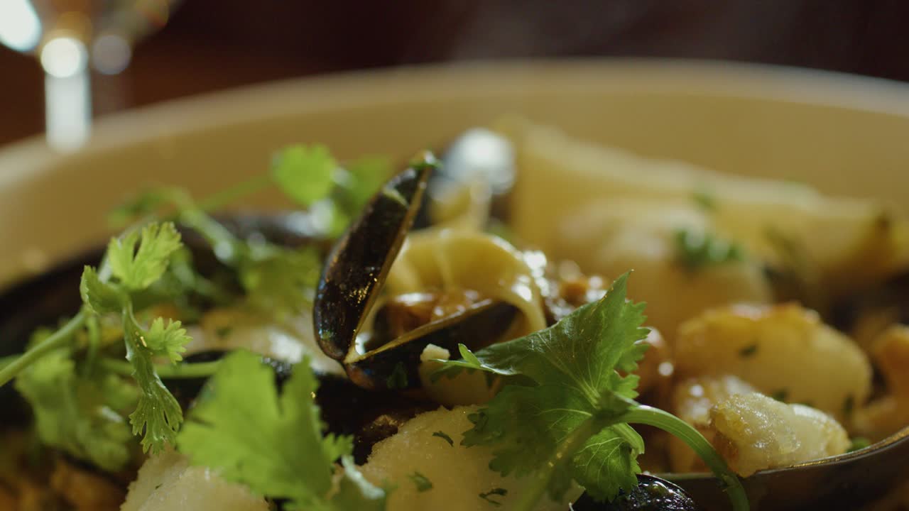 Close-up of hot seafood chowder with mussels, fish, and herbs, as a hand uses a spoon to scoop and eat in warm ambient lighting