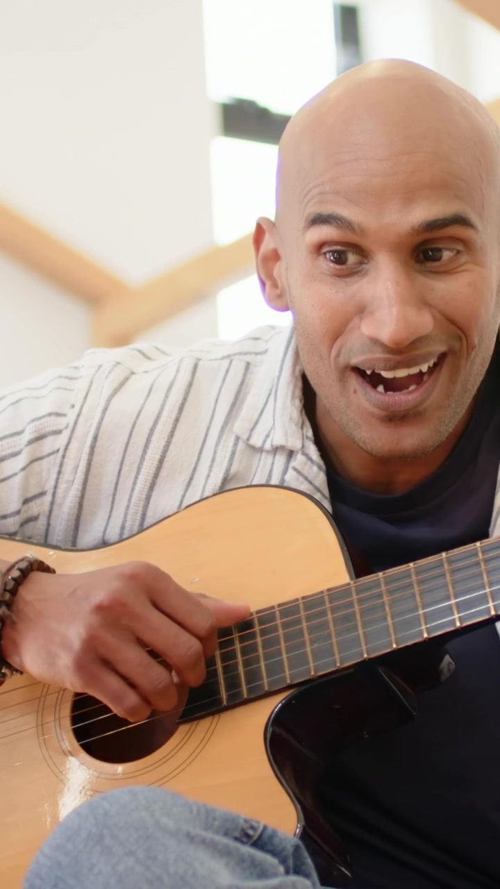 Video verticale: Uomo maturo che sistema la chitarra, suona e canta a casa, esercitandosi, con una camicia a righe.