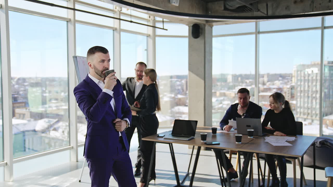 Middle-aged businessman standing in office, drinking coffee and thinking over something. Two teams working separately at the backdrop. Business staff brainstorm in sunlit office.