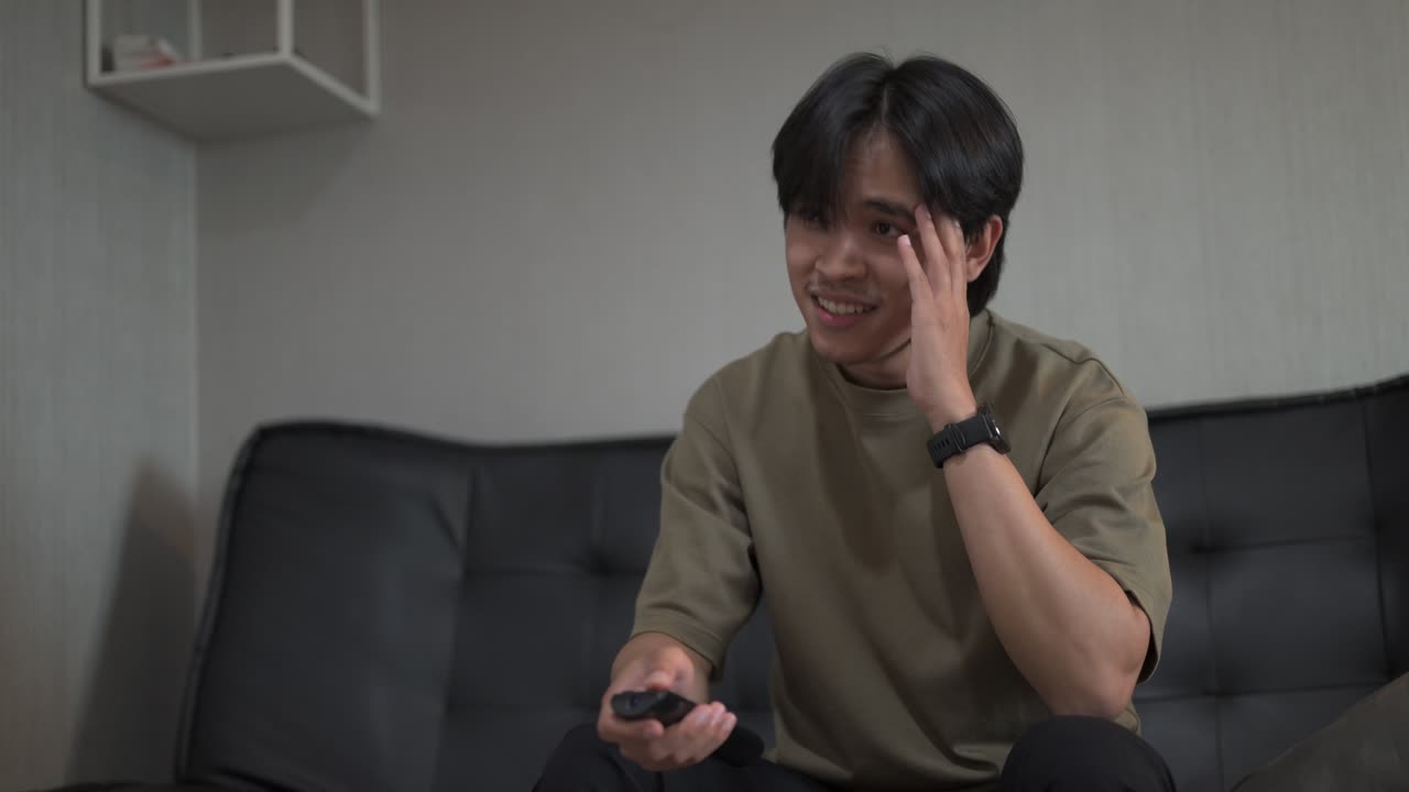 Handsome young Asian Thai man sitting at sofa indoors while looking television looking sad