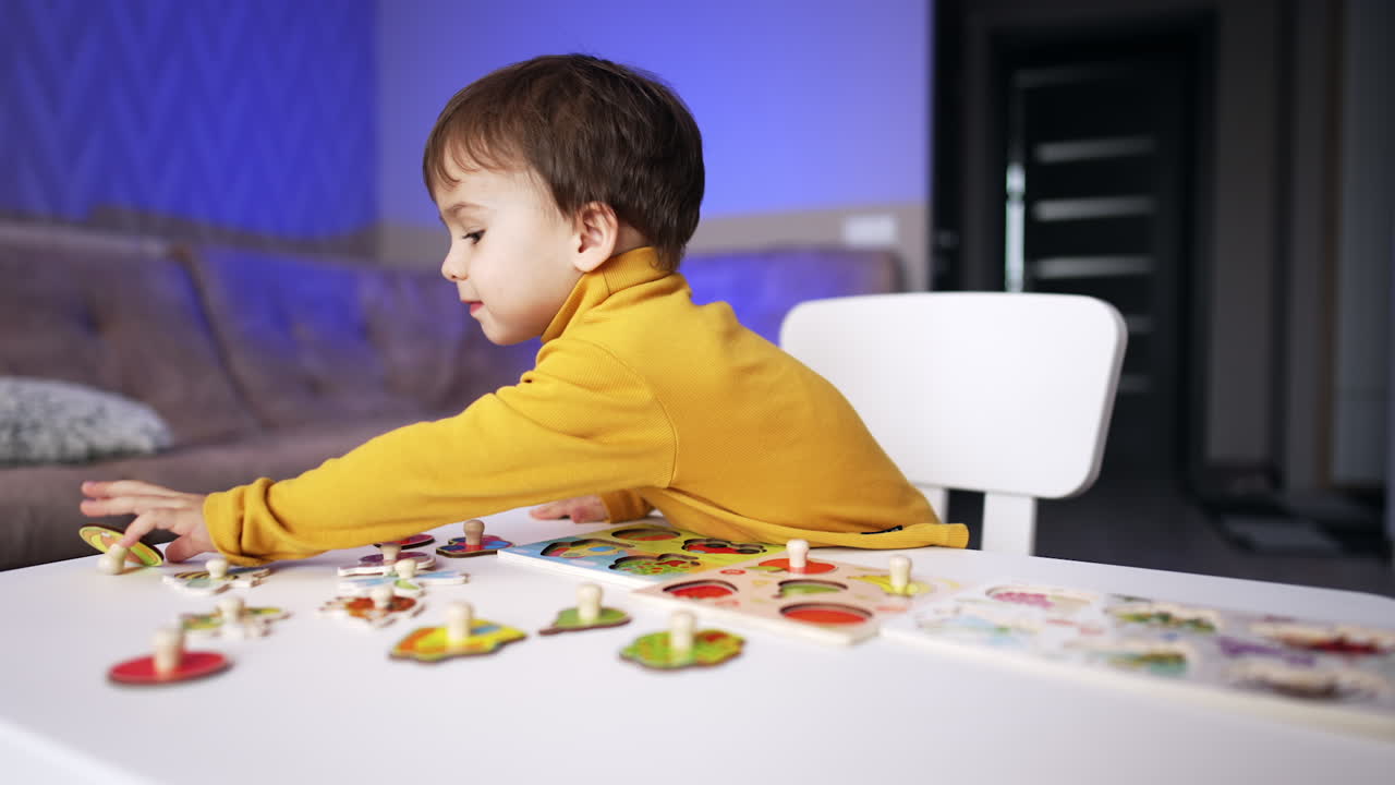 Lovely Caucasian baby wearing yellow sweater sits at desk. Toddler plays with puzzles at home. Blurred backdrop.