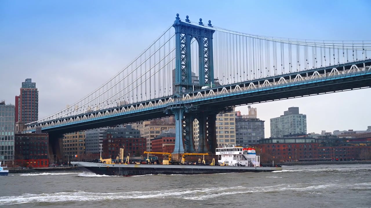 New York, USA, 26 maggio 2026: Ponte di Manhattan con una chiatta da carico che passa sotto. Campata sospesa in acciaio sopra l'East River con gli edifici di DUMBO