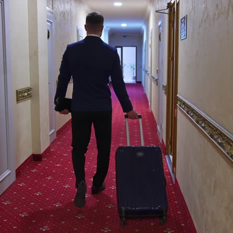 a man with a suitcase is walking down a hotel hallway. dark red carpet in a hotel hall. view from the back on a businessman in a trip