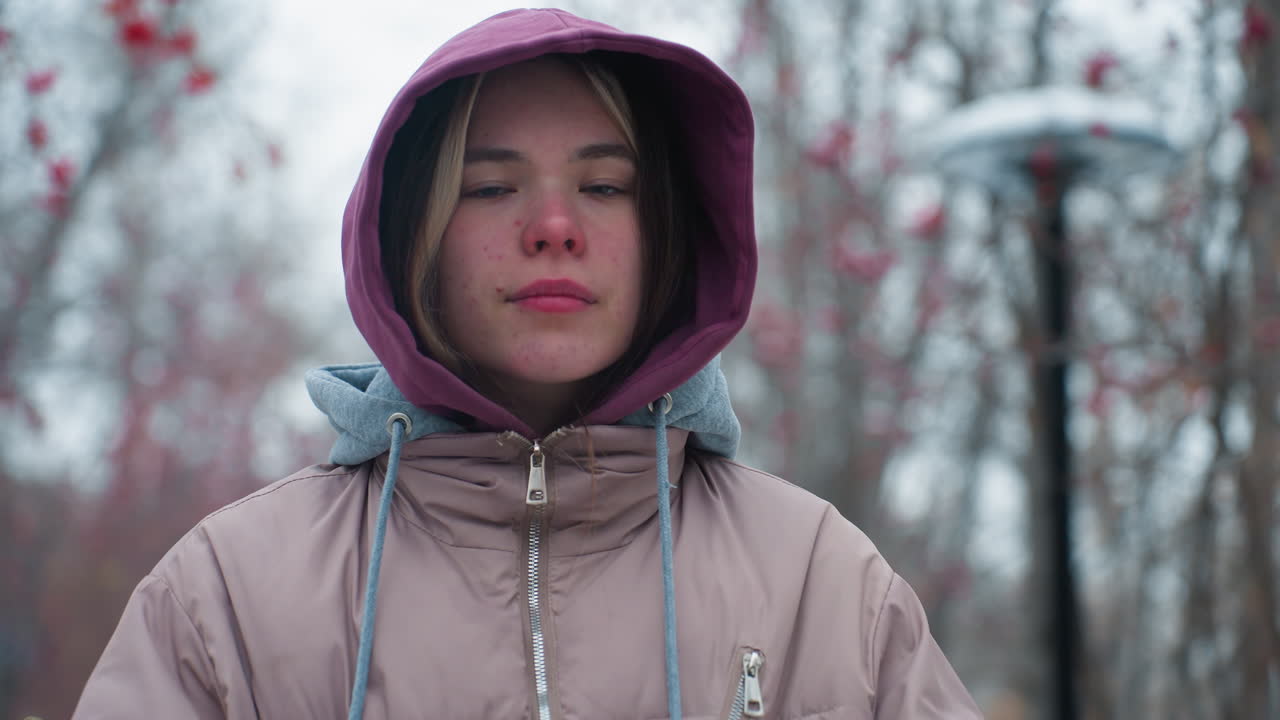 겨울 재을 입고 밖에서 자신감 있게 서있는 젊은 여자의 가까운 사진, 카메라에 대담한 시선으로 후디 모자를 조정, 나무와 가을 잎으로 흐릿한 배경