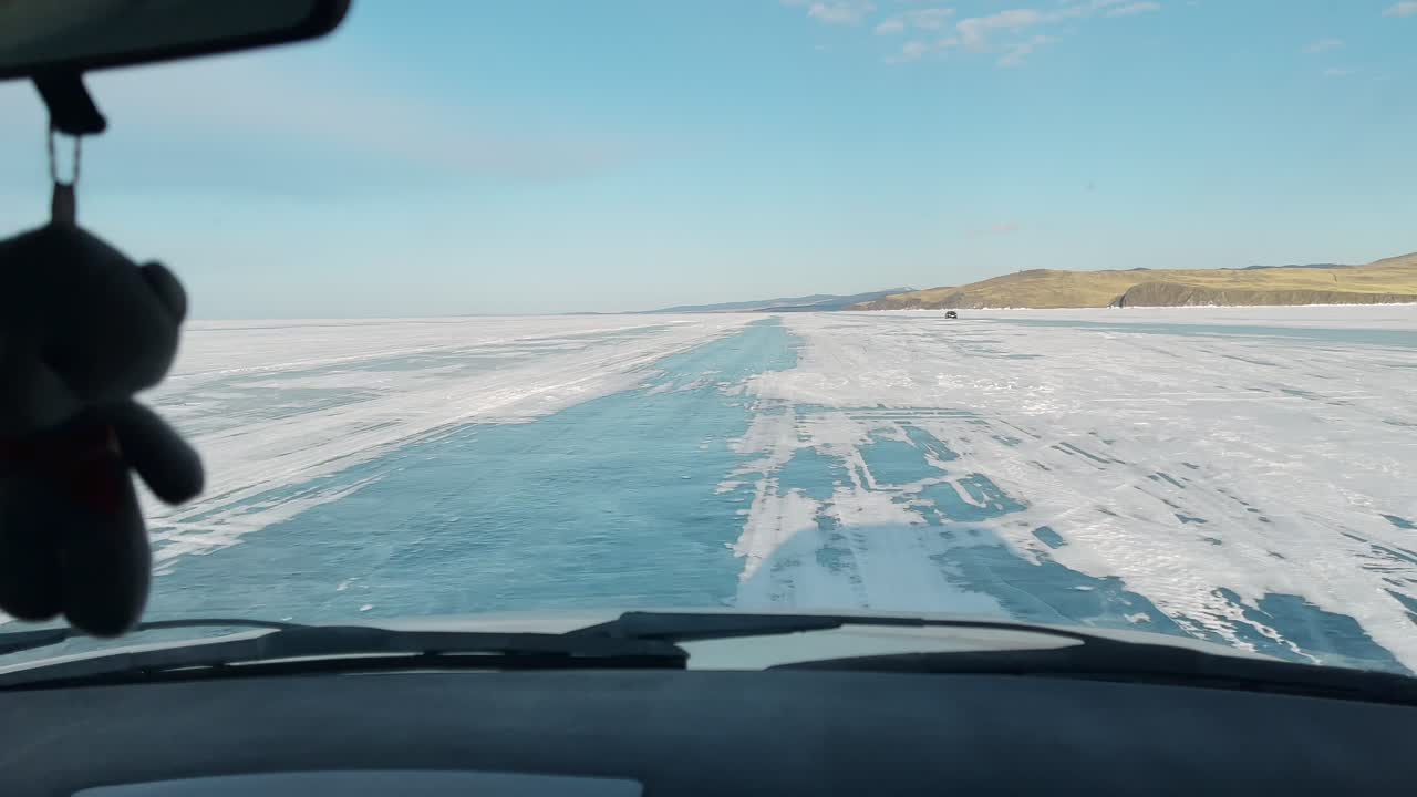 POV dall'interno di un veicolo che percorre una strada di ghiaccio blu attraverso il Lago Baikal ghiacciato in inverno, con superficie innevata e colline distanti sotto un cielo sereno, Russia.