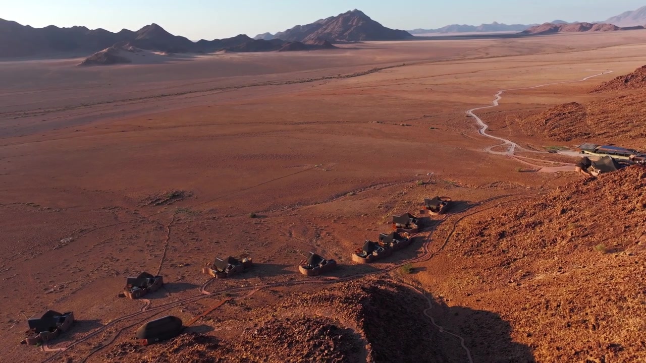 Vista aérea de drone de um alojamento no deserto com montanhas e uma planície infinita estendendo-se à distância do Namibe. Um refúgio remoto situado no coração da natureza selvagem do deserto.