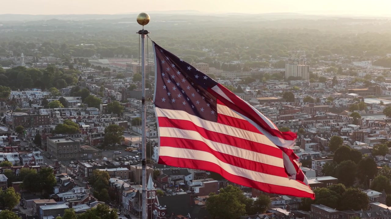 Amerika Birleşik Devletleri genelinde küçük kasaba çatılarının üzerinde Amerikan bayrağı dalgalanırken, beyaz kilise kulesi ve yoğun yerleşim sokakları sıcak altın rengi akşam güneşinin parıltısı altında yayılıyor. Havadan drone yakın çekim.