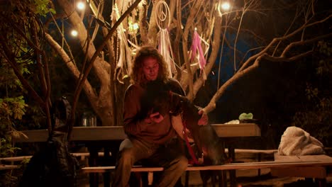 a curly-haired boy feeds a black dog on a bench near a campfire against a forest background. rest in the country house