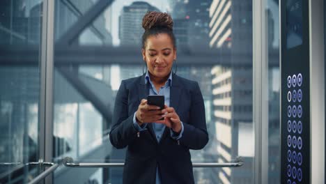 confident black adult female riding glass elevator to office in modern business center. successful african american manager using smartphone, write message, check social media and emails in a lift.