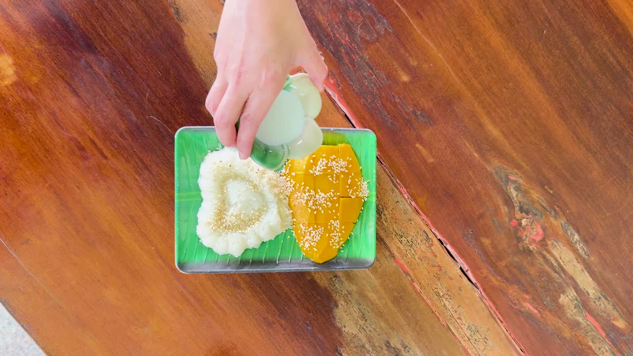 A hand pours coconut sauce over mango sticky rice on a wooden table. Bright lighting enhances the vibrant colors
