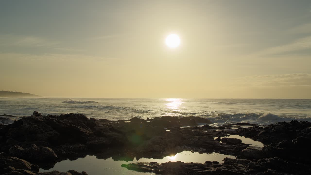 Waves crash against the rocks in the foreground, as the soft light of the early morning sun spreads across the horizon.