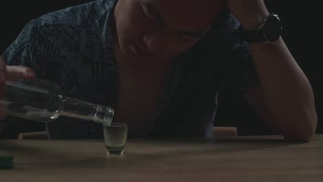 close up of drunk, depressed asian man pouring vodka in a shot glass before drinking in black background