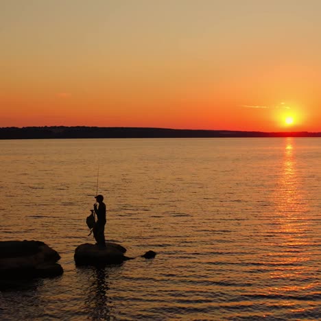 river at sunset. silhouette of a fisherman on stone. man going away from the lake after fishing on orange sky background in the evening.