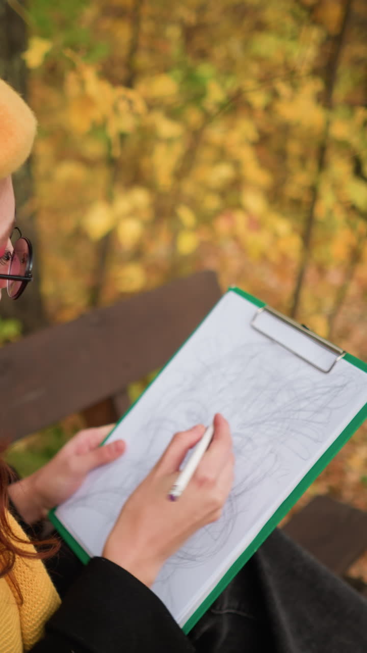 vista trasera de una joven con gafas de sol, boina amarilla y silenciador amarillo profundamente centrada en su dibujo, sentada en un banco en medio de un vibrante follaje de otoño