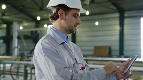 side view of engineer wearing a helmet and typing on the tablet in a big factory