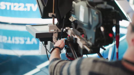 close-up of hand adjusting camera controls on professional broadcast camera rig in studio. teleprompter visible, operator managing setup and focusing on precise camera movements for filming