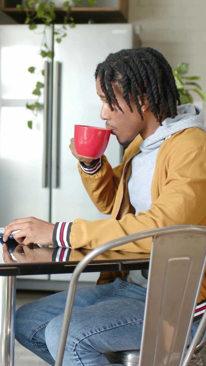 Vertical video: Working on computer, man in mustard coat sipping red mug using mouse in kitchen