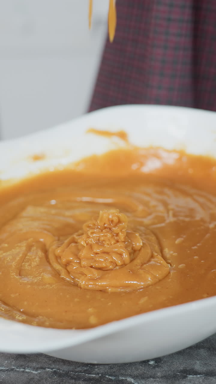 Baking process in action as whisk is lifted, causing thick batter to stretch and slowly drip into bowl, hands holding bowl and whisk are dusted with flour