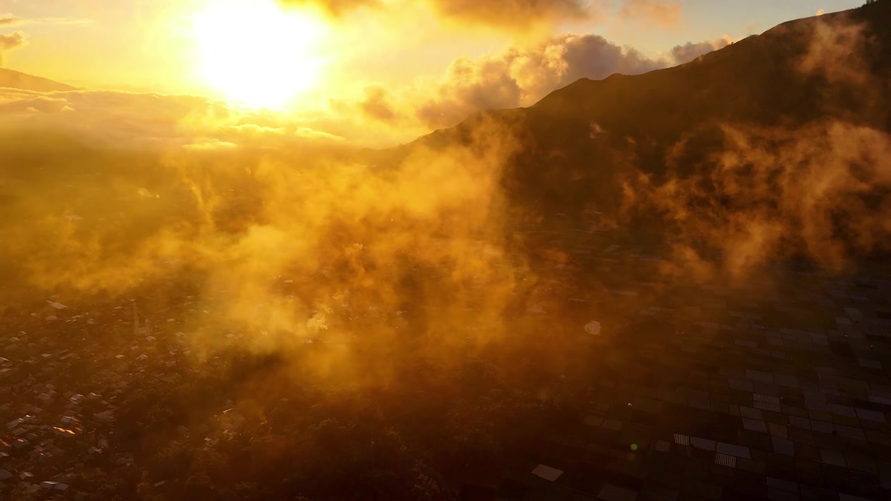 Ethereal golden mist drifts through Sembalun Valley at sunrise, aerial backdrop above dense home neighborhoods