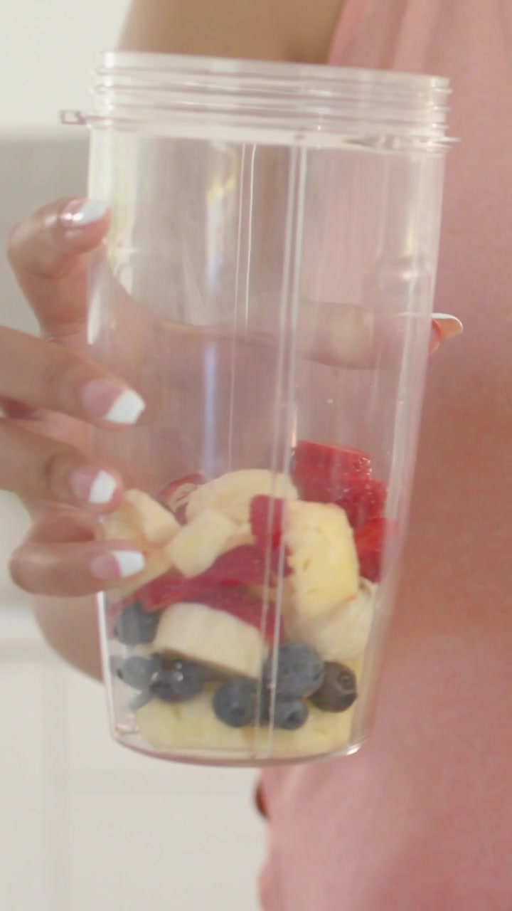 Video verticale: Donna con top rosa che inclina il bicchiere del frullatore con frutta in cucina, mentre prepara un frullato.