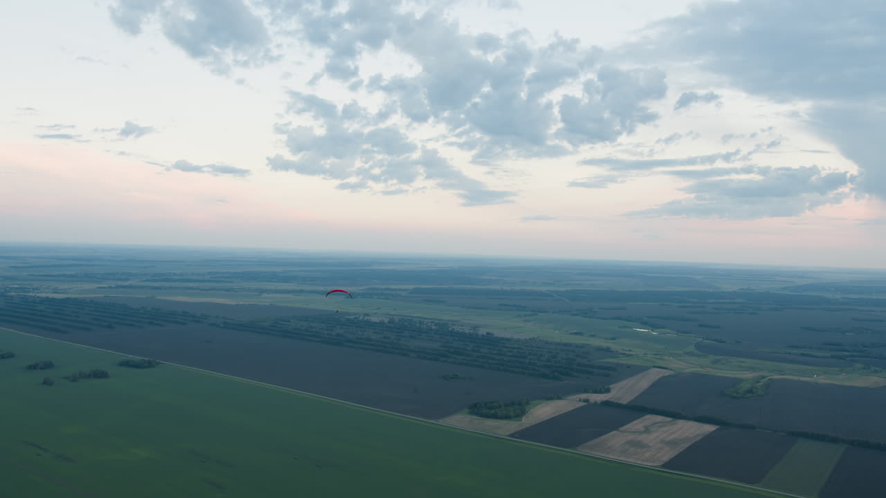Distant silhouette of pilot drifting paraglider under pastel dusk sky, clouds scattered overhead as canopy arcs above patchwork fields and tree lines in tranquil evening graceful flight