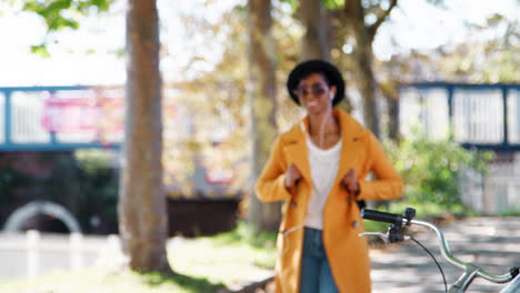 Una-Joven-Negra-De-Moda-Con-Sombrero,-Gafas-De-Sol,-Jeans-Azules-Y-Un-Abrigo-Amarillo-Caminando-Por-Una-Calle-Arbolada-En-Un-Día-Soleado-Escuchando-Música-Con-Auriculares,-Enfocada-En-Rack