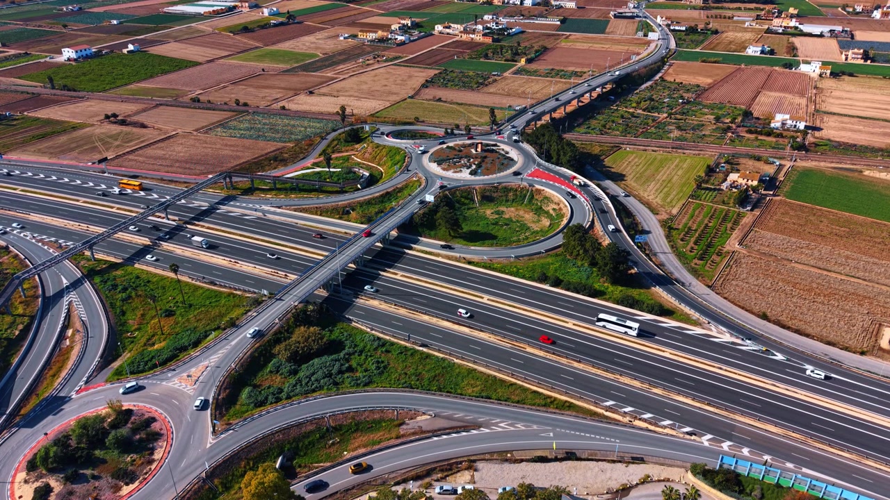 Aerial view of cloverleaf interchange surrounded by farmland, Spain. Oblique drone shot of looping ramps, fields and scattered houses.