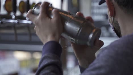 Slow-motion-shot-of-male-bartender-with-face-mask-shaking-cocktail-with-shaker-inside-bar